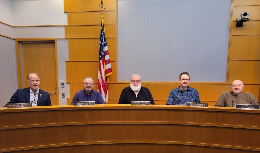 Five men sit behind a curved wooden desk in a council chamber with an American flag and a camera on the wall behind them. Each man has a nameplate in front of him. The group is smiling and facing forward.