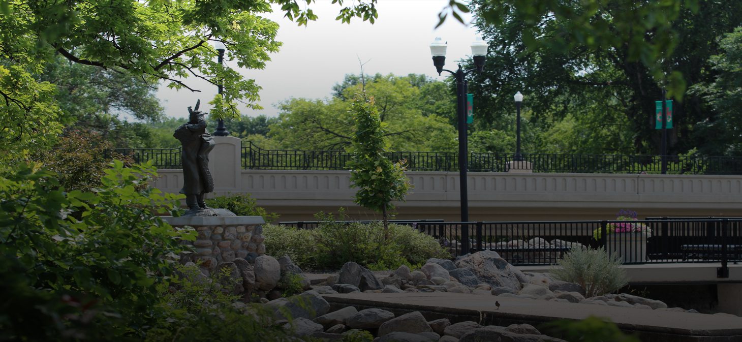 A bronze statue of a man stands on a stone pedestal surrounded by greenery and rocks near a bridge with black railings and streetlights on a bright, sunny day.