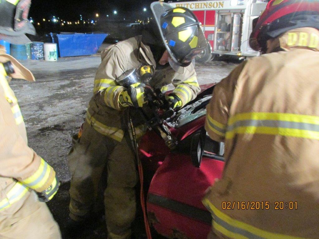 Three firefighters in full gear use hydraulic rescue tools on a red car during a night training exercise, with a fire truck visible in the background.
