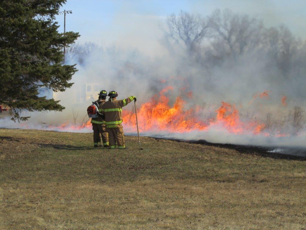 Two firefighters in full gear stand on dry grass, one pointing toward a line of fire and smoke burning through vegetation, with trees and hazy sky in the background.