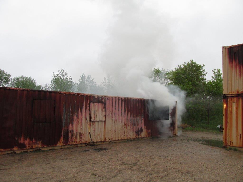 A rusted shipping container sits on dirt ground with smoke billowing out of one of its windows, surrounded by green trees under a cloudy sky.
