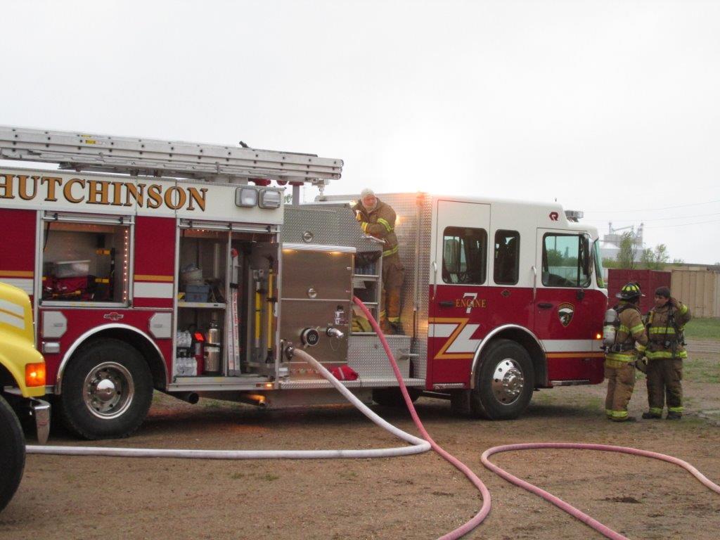 Three firefighters stand near a red fire truck labeled Hutchinson with hoses attached. Another firefighter is on the truck, preparing equipment. The background shows a building and cloudy sky.