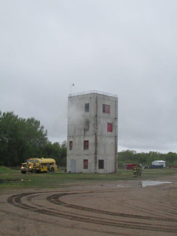 A four-story concrete training tower with red window covers emits light smoke from upper windows. A yellow fire truck is parked nearby, and the ground is muddy with tire tracks. Trees and cloudy sky are in the background.