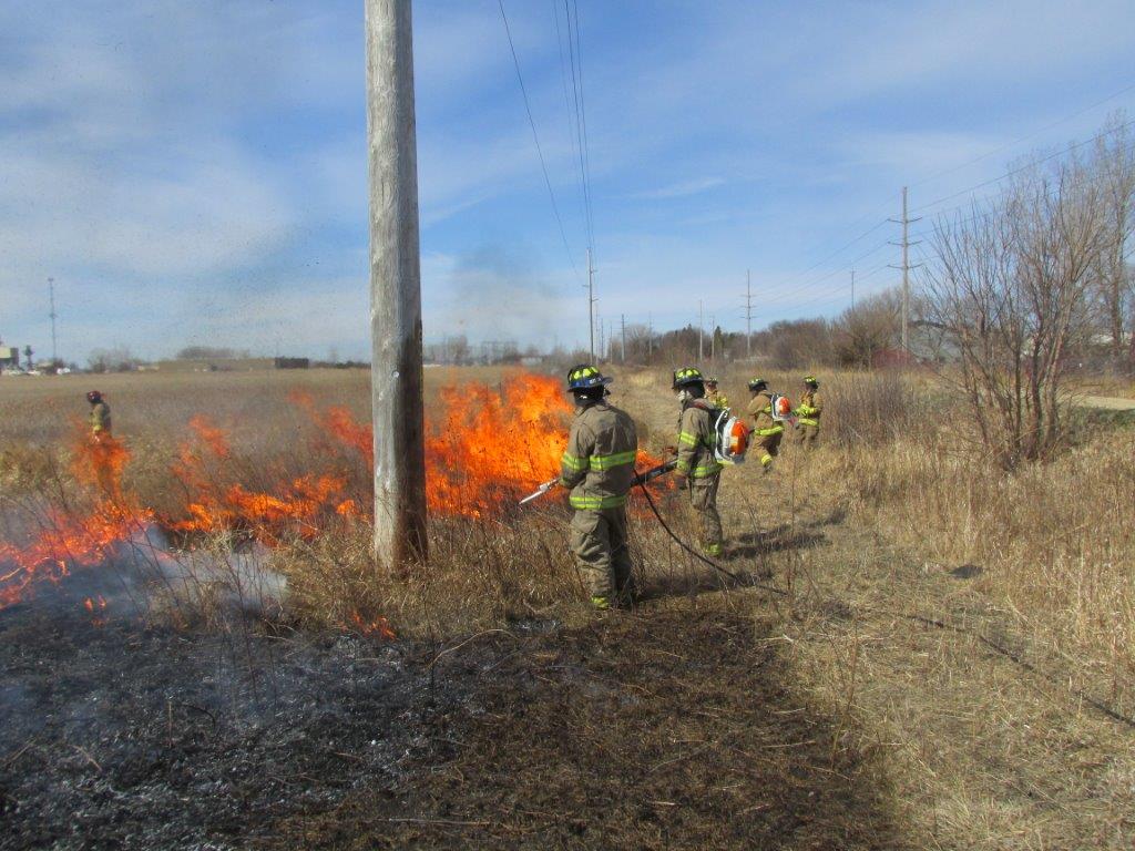 Firefighters in protective gear use hoses to extinguish a grass fire burning near a utility pole in a dry, open field under a partly cloudy sky.