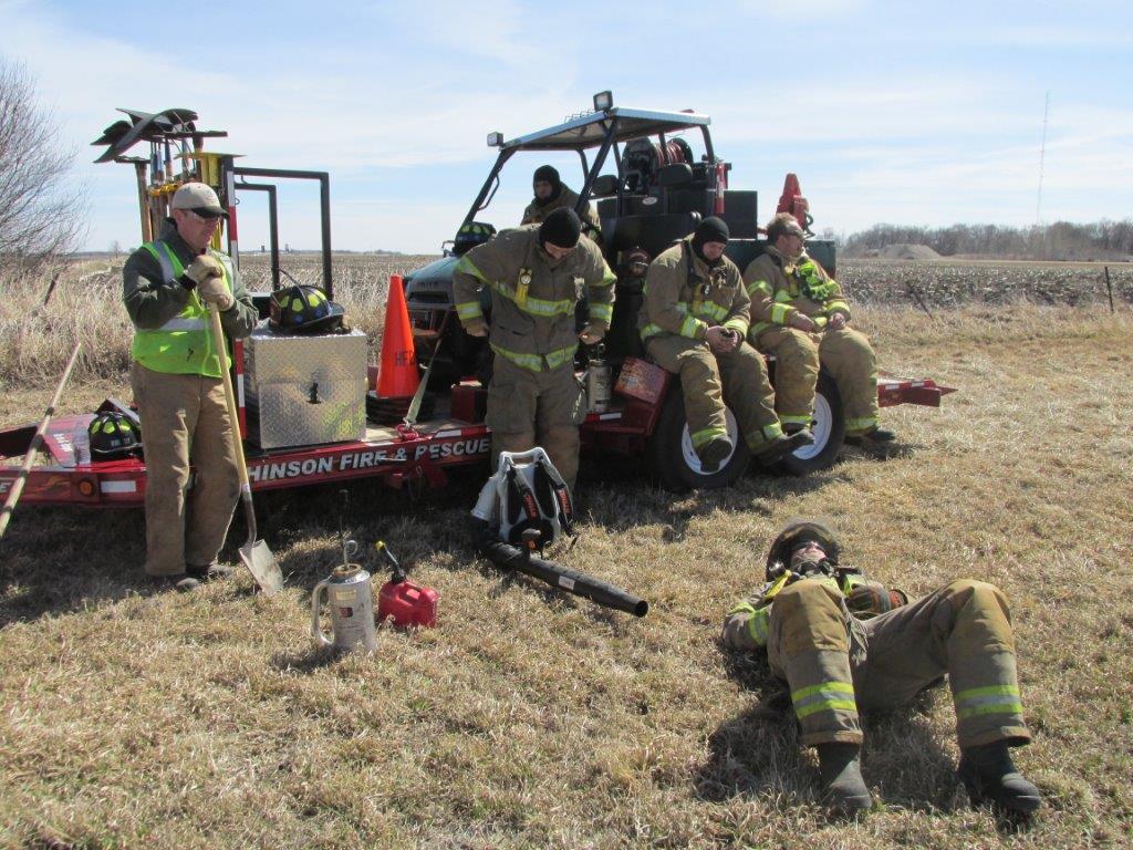 Five firefighters in full gear rest beside a rescue trailer on a grassy field, with one lying on the ground and others sitting or standing. Equipment like shovels and cones are visible. The sky is clear and sunny.