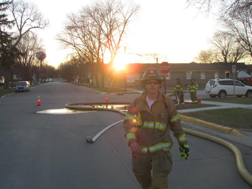A firefighter in full gear walks toward the camera on a residential street at sunset. Behind him, more firefighters stand near a large yellow hose, and orange cones block part of the road.