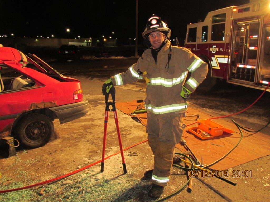 A firefighter in full gear stands next to a red car with a shattered window, holding a large bolt cutter beside firefighting equipment at night. A fire truck is visible in the background.